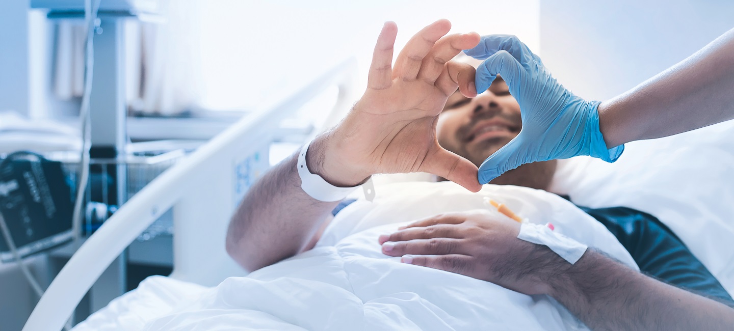 Patient im Klinikbett bildet mit der Hand einer Pflegekraft ein Herz.